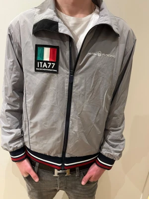 Sail Racing windbreaker grå - Snygg grå vindjacka från Sail Racing med ITA77-patch och italienska flaggan på bröstet. Storlek S