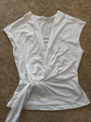 Vit omlottopp med knytband - Snygg vit omlottopp med knytband i midjan och v-ringad hals. Toppen är ärmlös och har en stilren, enkel design som passar till många tillfällen. Finns ingen lapp tyvärr