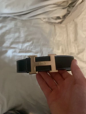 Svart skärp med guldfärgat H-spänne - Snyggt svart skärp med ikoniskt guldfärgat H-spänne. Skärpet har vita kontrastsömmar längs kanterna och är gjort i slätt skinn. Perfekt accessoar för att lyfta vilken outfit som helst.