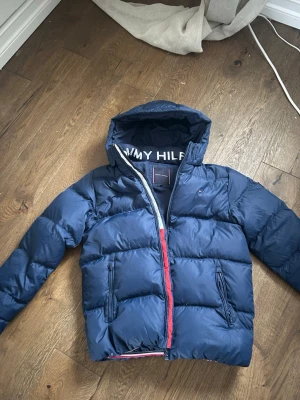 Mörkblå pufferjacka från Tommy Hilfiger - Snygg mörkblå pufferjacka från Tommy Hilfiger med huva och dragkedja. Jackan har två framfickor med dragkedja och logotyp på insidan av huvan. Perfekt för kalla vinterdagar och har en klassisk quiltad design. Jackan är storlek 164