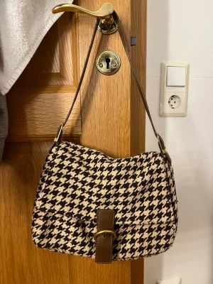 Houndstooth handväska med brunt spänne - Snygg handväska i klassiskt svartvitt hundtandsmönster med brunt justerbart axelband och dekorativt spänne i guld. Väskan har en mjuk, tygklädd yta och en rektangulär form med lock. Perfekt accessoar för att lyfta din outfit.