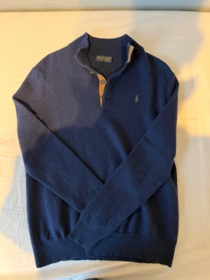 Mörkblå stickad tröja Polo Ralph Lauren Golf - Mörkblå stickad tröja från Polo Golf Ralph Lauren med knappslå i kontrasterande beige och små bruna knappar. Tröjan har lång ärm, ribbade muddar och en liten grön logga broderad på bröstet. Perfekt för en stilren och avslappnad look. Varm stickad tröja. Perfekt nu under vintern. Storlek XS eventuell S. Inga synliga tecken på användning Pris kan diskuteras
