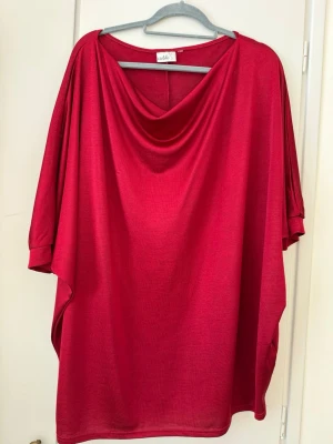 Röd loose topp med vid hals Isolde  - Snygg röd topp från Isolde med lös passform och vid, draperad halsringning. Toppen har korta, vida ärmar och är tillverkad i ett mjukt, tunt material som faller fint. Perfekt för dig som gillar en avslappnad och trendig stil.