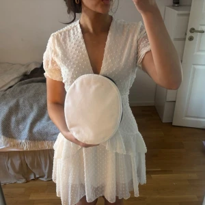 Vit kortklänning med volang och prickar - Säljer en söt vit kortklänning med v-ringning, volangdetaljer och prickigt mönster. Klänningen har korta ärmar och omlottliknande överdel. Perfekt för sommarens alla tillfällen!