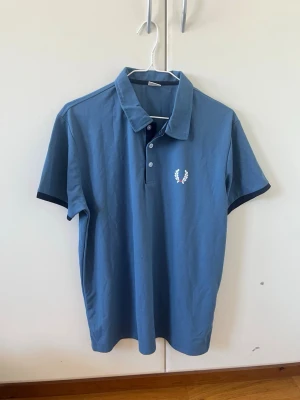 Blå pikétröja  - Snygg blå pikétröja med klassisk krage och knappar framtill. Tröjan har den ikoniska vita loggan broderad på bröstet och mörkare detaljer vid ärmsluten. Perfekt för en avslappnad men stilren look.