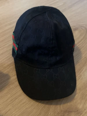 Svart Gucci keps med GG-mönster - Snygg svart keps från Gucci med klassiskt GG-monogram över hela kepsen. På sidorna finns de ikoniska gröna och röda ränderna. Kepsen har böjd skärm och är tillverkad i ett slitstarkt tygmaterial. Perfekt för dig som vill ha en exklusiv och trendig accessoar.