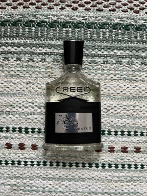 Creed Aventus Eau de Parfum - Creed Aventus Eau de Parfum med elegant transparent glasflaska, svart etikett och silverdetaljer. Flaskan har en svart plastkork och en stilren, exklusiv design. Märkets logga och Aventus-etiketten syns tydligt på framsidan. Förpackningen är svart med silverdetaljer.