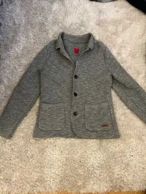 Grå kofta med knappar från S.oliver - Snygg gråmelerad kofta från S.oliver med tre stora knappar framtill och två rymliga fickor. Koftan har en klassisk krage och är tillverkad i mjukt stickat material. Perfekt för lager på lager och enkel att matcha med olika outfits. Aldrig använd! Strlk xs! Rekommenderar att vara runt 155-165 ish! 