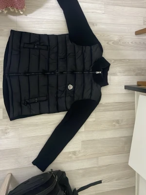 Svart dunväst från Moncler - Snygg svart dunväst från Moncler med quiltad design och två framfickor med knappstängning. Västen har en hög krage och dragkedja framtill samt det klassiska Moncler-emblemet på bröstet. Perfekt för lager-på-lager och streetstyle.