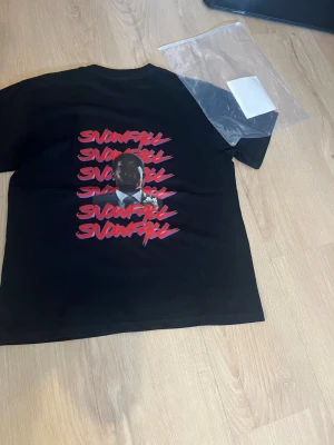 Svart SNOWFALL t-shirt med tryck - Svart t-shirt från SNOWFALL med röd och vit text på ryggen och ett stort tryck av ett ansikte. På bröstet finns en liten röd ros och SNOWFALL-logga. Klassisk passform och rund hals. Perfekt för dig som gillar streetwear och snygga prints.