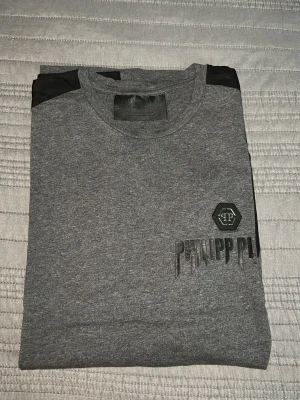 Grå Philipp Plein t-shirt med svarta detaljer - Snygg grå t-shirt från Philipp Plein med svarta paneler längs axlar och sidor. T-shirten har rund halsringning och tryckt logga på bröstet. Perfekt för dig som gillar streetwear och vill sticka ut lite extra.