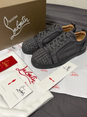 Grå Louboutin sneakers -  gråa Louboutins med spikar och klassisk röd sula. Skorna har en låg profil, är gjorda i mocka och har gråa skosnören. Perfekt för dig som vill sticka ut med exklusiva detaljer och lyxig design.