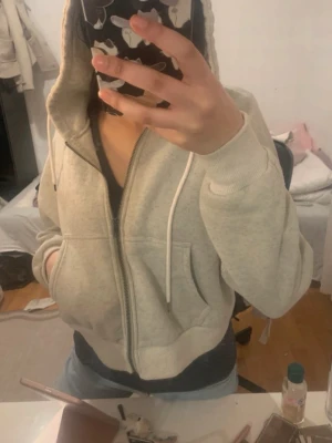 Ljusgrå zip hoodie med huva - Säljer en ljusgrå hoodie med dragkedja och huva. Hoodien har vita dragsnören, fickor framtill och ribbade muddar vid ärmslut och nederkant. Perfekt för en avslappnad och chill stil. Materialet känns mjukt och bekvämt.