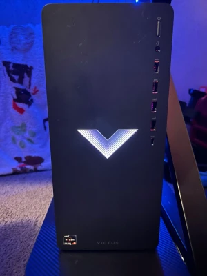 HP Victus stationär gamingdator - Säljer en svart HP Victus stationär dator med stilren design och belyst V-logga på fronten. Utrustad med flera USB-portar och AMD Ryzen 7-processor. Perfekt för gaming och multitasking. Kompakt och modern låda i metall och plast.