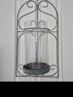 Chabby chick mysig Väggljusstake i metall med glasinsats! - Snygg vägglykta med grå metallram i dekorativt mönster och cylinderformad glascylinder. Perfekt för blockljus eller värmeljus. Ger mysig stämning och passar bra som inredningsdetalj. Även Perfekt för att skapa en mysig atmosfär i hemmet. Lätt att hänga upp på väggen. Ca 35-40 cm hög. Svart/silver ljus ingår. Fint med vitt blockljus till.  Mycket fint skick! Djur och rökfritt hem