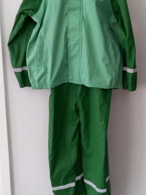 Barn - Grön regnställ med reflexdetaljer från Exani  - Storlek 122 - Regnjacka i två nyanser av grönt med reflexband på ärmarna. Jackan har dragkedja, tryckknappar och två fickor. Tillverkad i vattentätt syntetmaterial och extra knappar medföljer. Perfekt för regniga dagar. Barnstorlek 122 motsvarar en längd på  cm och passar vanligtvis barn i åldern 6–7 år. Storleken är avsedd för barn som är cirka 117–122 cm långa. Midjevidden för denna storlek ligger ofta kring 57–58,5 cm. Den passar oftast i övergången från förskoleklass till lågstadiet.