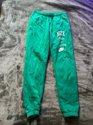 Gröna mjukisbyxor från Nike - Snygga gröna mjukisbyxor från Nike med tryckta loggor på vänster ben. Byxorna har resår i midjan och vid bensluten samt sidofickor. Perfekta för chill dagar eller träning. Tillverkade i mjukt material för maximal komfort.