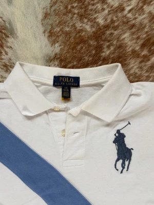 Vit pikétröja från Polo Ralph Lauren - Vit pikétröja från Polo Ralph Lauren med blå diagonal rand och stor broderad logga på bröstet. Klassisk krage, knappar framtill och blå siffra 3 på ärmen. Perfekt för en sportig och stilren look. Köpt på NK. Nyskick 9,5/10. Perfekt till våren. Passar S/xs. Nypris 1200. Bara att höra av sig vid frågor//Carl