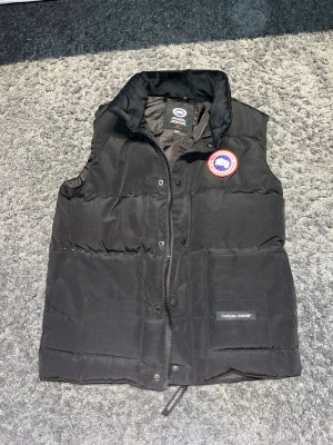 Svart dunväst från Canada Goose - Snygg svart dunväst från Canada Goose med klassisk logga på bröstet och hög krage. Västen har dragkedja och knappar framtill samt två stora fickor. Perfekt för lager-på-lager och håller dig varm under kyliga dagar.