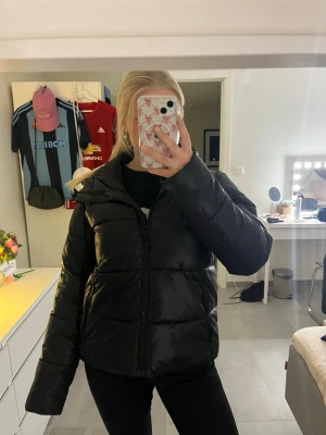 Svart pufferjacka med huva - Säljer en svart pufferjacka med dragkedja och stor huva. Jackan har en boxig passform och breda kanaler som ger en riktigt puffig look. Perfekt för kalla dagar och enkel att styla till allt. Materialet är glansigt och ger en modern vibe.