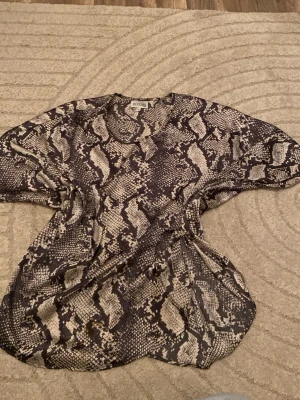 Ormmönstrad blus från By Jolima - Snygg blus från By Jolima med trendigt ormmönster i svart och beige. Modellen är lös och luftig med korta ärmar och rund halsringning. Perfekt för dig som vill sticka ut med ett djurmönstrat plagg. Materialet känns lätt och bekvämt mot huden.