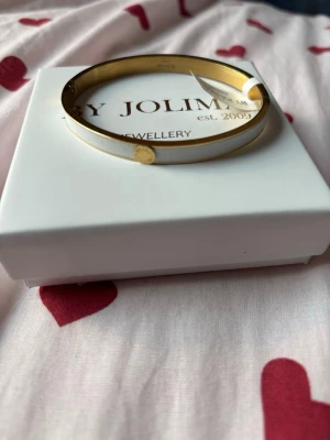 Guldigt armband från By Jolima - Snyggt armband från By Jolima i guldfärgad metall med vit emaljdetalj och rund guldfärgad dekoration. Levereras i original ask. Aldrig använd🥰
