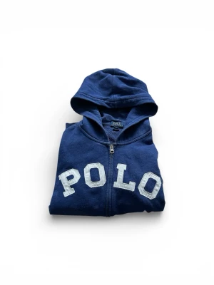 Mörkblå zip hoodie från Polo Ralph Lauren - Snygg blå hoodie med dragkedja från Polo Ralph Lauren. Stora vita bokstäver 'POLO' på bröstet, huva och praktiska fickor framtill. Perfekt för en avslappnad och cool look. Materialet är mjukt och bekvämt, troligtvis bomullsblandning.