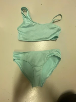 Ljusblå ribbad bikini med enkel axel - Snygg ljusblå bikini med ribbad struktur och enkel axel på toppen. Både överdel och underdel har en stilren och sportig look. Perfekt för strandhäng eller poolparty!