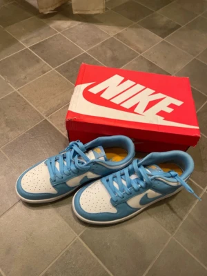 Nike Dunk Low Retro blå/vit - Nike Dunk Low Retro sneakers i blått och vitt med klassisk låg siluett. Skorna har vita läderpaneler, blå detaljer och matchande blå snören. Sulan är vit med blå yttersula och innersulan har gul Nike-logga. Perfekt för dig som gillar streetwear och snygg design.