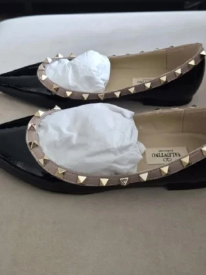 Valentino Rockstud ballerinaskor svart - Snygga svarta ballerinaskor från Valentino med glansig finish och beige kant dekorerad med ikoniska guldfärgade nitar. Klassisk spetsig tå och låg klack. Tillverkade i läder för en exklusiv känsla och look.