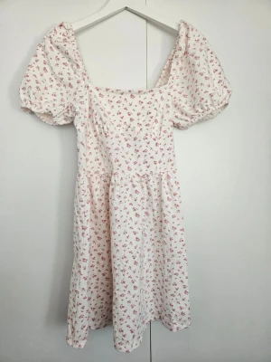 Vit klänning med rosa blommor - Supersöt vit klänning med små rosa blommor och puffiga korta ärmar. Klänningen har en fyrkantig ringning och är gjord i mjuk bomull, perfekt för en fräsch och romantisk look.