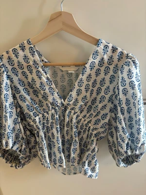 Vit blus med blått mönster H&M - Supersöt vit blus från H&M med blått blommigt mönster. Blusen har v-ringning, puffiga trekvartsärmar med resår vid ärmslut och smockad midja som ger en snygg siluett. Tillverkad i ett lätt och luftigt material, perfekt för sommardagar. Väldigt trendig!! Aldrig använd.