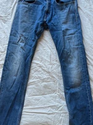 Blå raka replay jeans med slitningar - Säljer ett par klassiska blå replay jeans med raka ben och diskreta slitningar på framsidan. Jeansen har fem fickor och är gjorda i denim med snygg tvätt. Perfekt för dig som gillar en enkel och tidlös look.