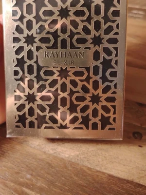 Rayhaan Elixir Parfym - Snygg parfym från Rayhaan med namnet Elixir. Flaskan har ett unikt geometriskt mönster i guld och svart som ger en lyxig känsla. Förpackningen är fyrkantig och har en elegant metallisk finish som verkligen sticker ut i badrumshyllan.