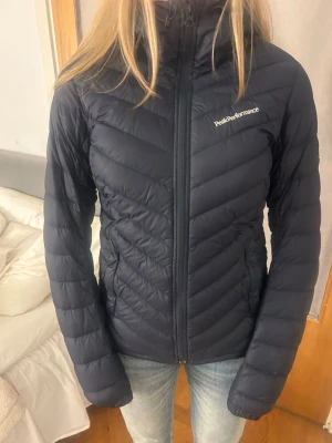 Svart/ mörkblå  dunjacka från Peak Performance - Snygg svart dunjacka från Peak Performance med huva och dragkedja framtill. Jackan har ett quiltat mönster och logga på bröstet. Perfekt för kalla vinterdagar och riktigt skön att bära. 