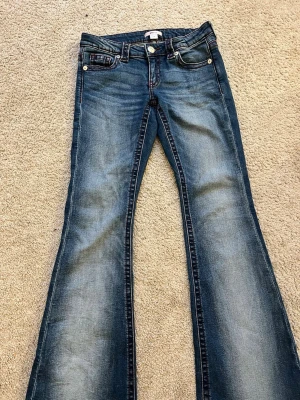 Blå bootcut jeans från Only - Snygga blå bootcut jeans från Only med slitningar och kontrastsömmar. Klassisk femficksmodell med knappgylf och bälteshällor. Jeansen har en cool tvättad look och är perfekta för en avslappnad stil. Materialet är bomull med lite stretch för extra komfort.