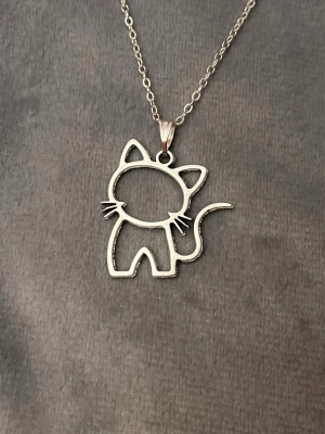 Katt halsband silver imitation  - Sött halsband med en berlock i form av en siluett av en katt. Kedjan är i silverimitation och justerbar 40-45 cm. Berlocken är ca 2,4 x 2,6 cm.  