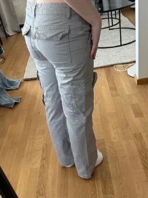 Ljusgrå cargobyxor med fickor - Säljer ett par ljusgrå cargobyxor med raka ben och flera praktiska fickor på sidorna och bak. Byxorna har bälteshällor och stängs med dragkedja och knapp. Perfekta för en avslappnad och trendig look.