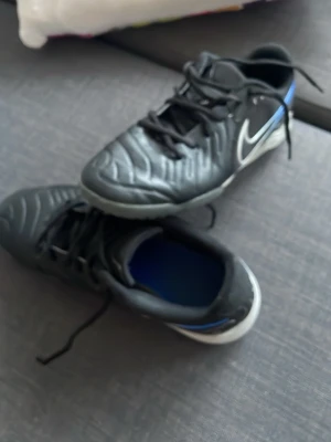 Nike svart/blå fotbollssko inomhus pris kan diskuteras  - Säljer ett par svarta Nike fotbollsskor för inomhusbruk med vit och blå detalj samt vit sula. Skorna har snörning, låg profil och är tillverkade i syntetmaterial med meshpaneler för bra ventilation. Perfekta för futsal eller inomhusträning.