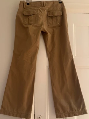 Beiga bootcut jeans med coola fickor - Snygga beiga jeans med bootcut-passform och två stora fickor med lock bak. Klassisk design och dubbla knappar fram. Bra skick och kvalitet 100% bomull