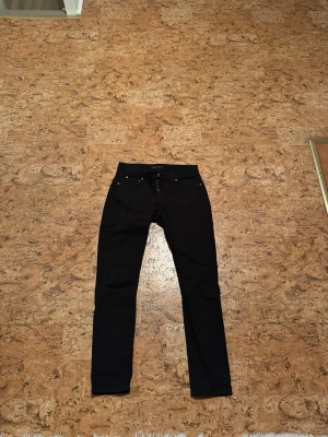 Nudie jeans - Säljer ett par svarta  jeans med klassisk femficksdesign och smal passform. Jeansen har bälteshällor och knappgylf. Perfekta för dig som gillar en stilren och enkel look. 