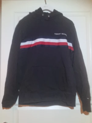 Svart hoodie från Tommy Hilfiger - Svart hoodie med huva från Tommy Hilfiger. Framsidan har ett broderat logotyptryck och en bred rand i blått, vitt och rött över bröstet. Klassisk passform med känguruficka och ribbade muddar. Perfekt för en avslappnad och cool look.