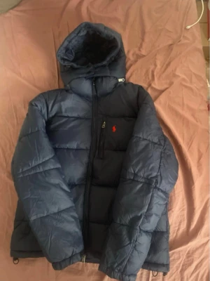 Mörk blå pufferjacka från Polo Ralph Lauren - Mörk blå pufferjacka från Polo Ralph Lauren med huva och två sidofickor med dragkedja. Jackan har en röd Polo-logga broderad på bröstet och snygga quiltade paneler. Materialet är syntet och jackan stängs med dragkedja framtill. Kvitto finns. Pris går att diskuteras vid ett snabbt köp.