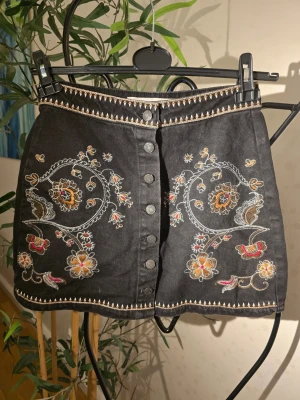 Svart broderad kortkjol med knappar - Säljer en svart kortkjol med broderade blommor i beige, brunt och rött. Kjolen har dekorativa knappar framtill och detaljerade sömmar längs midja och nederkant. Perfekt för dig som gillar unika detaljer och boho-vibe.