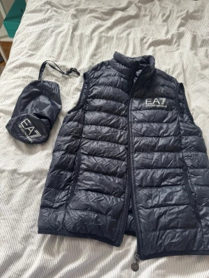 Svart dunväst från EA7 Emporio Armani aldrig använd typ - Snygg svart dunväst från EA7 Emporio Armani med quiltad design och hög krage. Västen har dragkedja framtill och logga på bröstet. Kommer med tillhörande förvaringspåse, perfekt att ta med på språng.