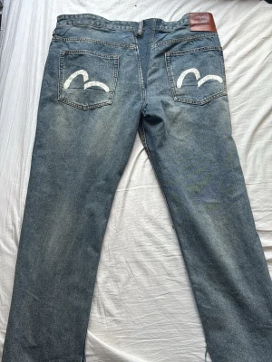Blå jeans från Evisu med målad bakficka - Säljer ett par klassiska blå jeans från Evisu med raka ben och ikoniska vita målad design på bakfickorna. Jeansen har fem fickor, knappgylf och en brun läderpatch på linningen. Materialet är slitstark denim i bomull.