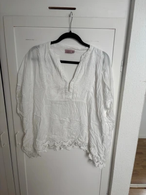 Vit boho-blus med spetsdetaljer - Säljer en luftig vit blus från Indiska med v-ringning, smockad överdel och fina spetsdetaljer längs nederkanten. Blusen har långa, vida ärmar och är tillverkad i ett tunt, mjukt tyg som ger en avslappnad boho-känsla. 