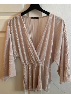 Beige meshblus från Gina Tricot - Trendig beige meshblus från Gina Tricot med omlott-skärning och djup V-ringning. Blusen har vida trekvartsärmar och resår i midjan för en snygg siluett. Tillverkad i ett transparent, ribbat meshmaterial som ger en luftig och modern känsla.