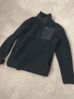 Svart fleecejacka med bröstficka - Mysig svart fleecejacka med halv dragkedja och en praktisk bröstficka med dragkedja. Jackan har hög krage och två sidofickor, perfekt för kalla dagar. Materialet är mjukt och värmande, och modellen är enkel men stilren.Storlek S men kan funka till M också!