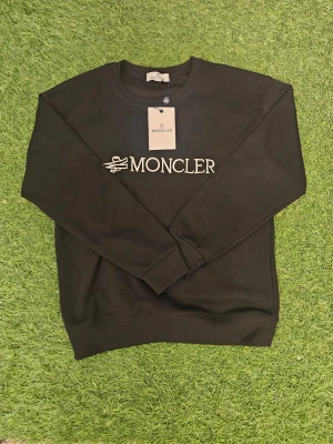 Svart stickad tröja från Moncler - Snygg svart stickad tröja från Moncler med rund halsringning och vit broderad logga på bröstet. Tröjan har ribbade muddar vid ärmslut och nederkant, vilket ger en clean och stilren look. Perfekt för dig som gillar exklusiva märken och enkel design.